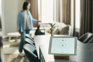 A smart home security tablet sits on a kitchen counter displaying a locked icon, while a woman in the background walks through a bright, modern living room.