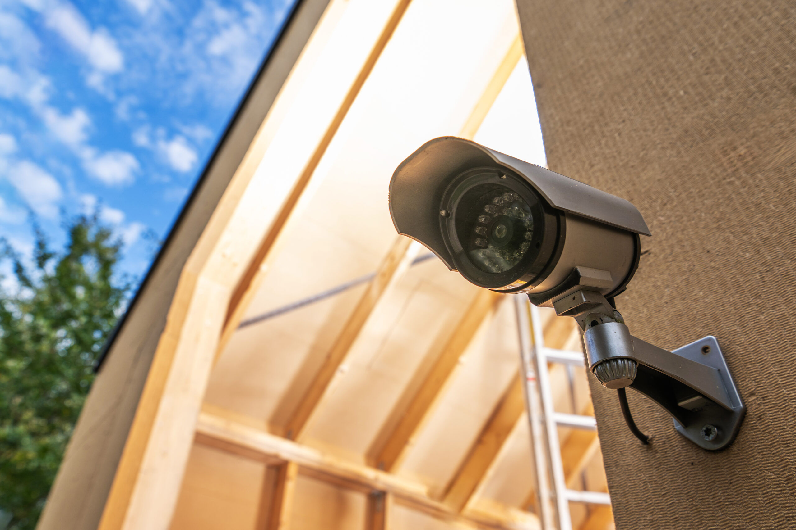 Construction Site Monitoring Using CCTV A mounted outdoor security camera on the side of a building under construction, with exposed beams and a blue sky in the background.