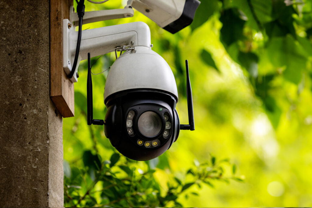 A dome-style outdoor security camera with dual antennas mounted on a concrete post, surrounded by bright green foliage.