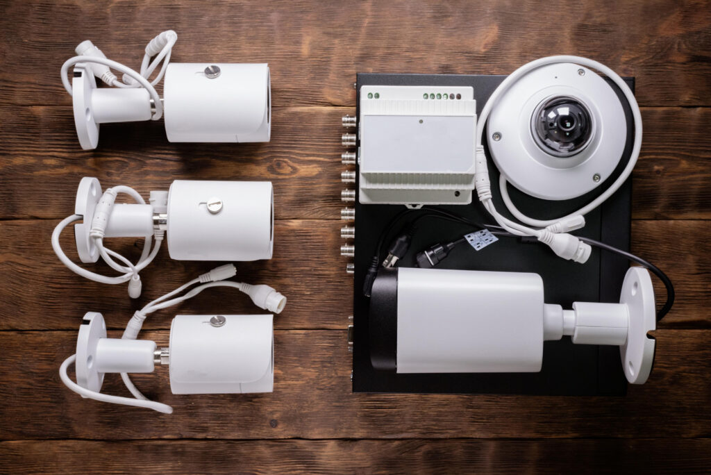A set of white security cameras and accessories laid out on a wooden surface, including bullet cameras, a dome camera, cables, and a DVR unit.