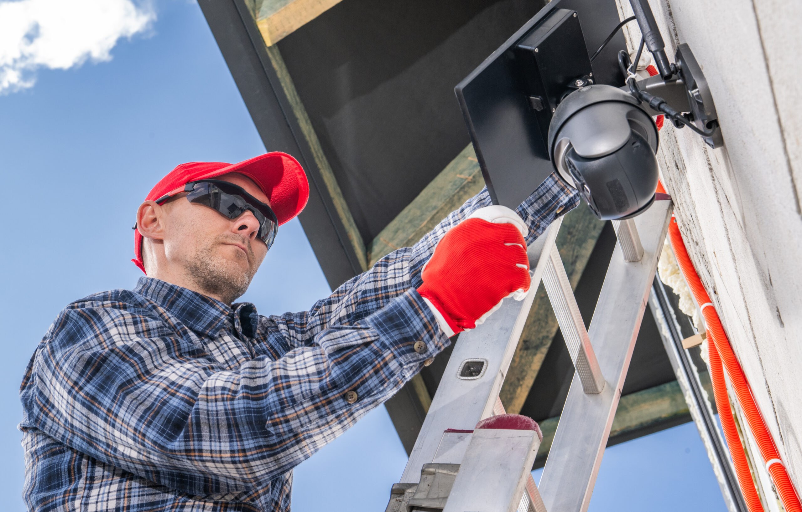Technician Installs Exterior Security Camera on a Residential Building During Bright Day in a Suburban Neighborhood A technician wearing a red cap, safety glasses, and gloves stands on a ladder installing an outdoor security camera on the exterior of a building.
