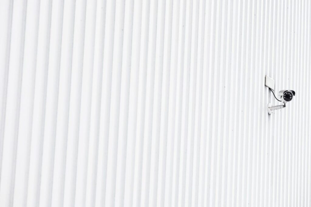 A security surveillance camera mounted on a white corrugated metal wall, angled outward to monitor the area, with clean lines and ample negative space.