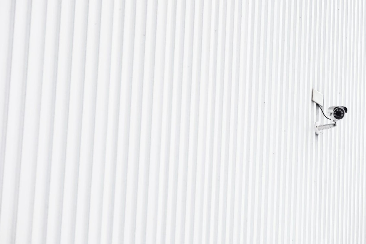 A security surveillance camera mounted on a white corrugated metal wall, angled outward to monitor the area, with clean lines and ample negative space.