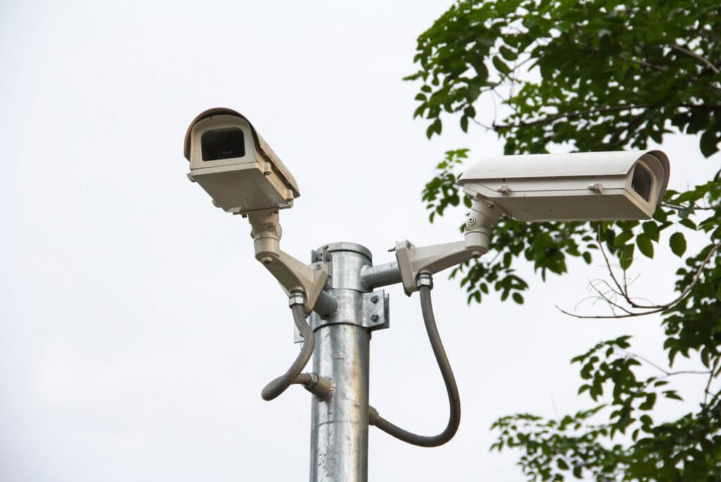 Outdoor security cameras mounted on a metal pole near trees in an urban area.